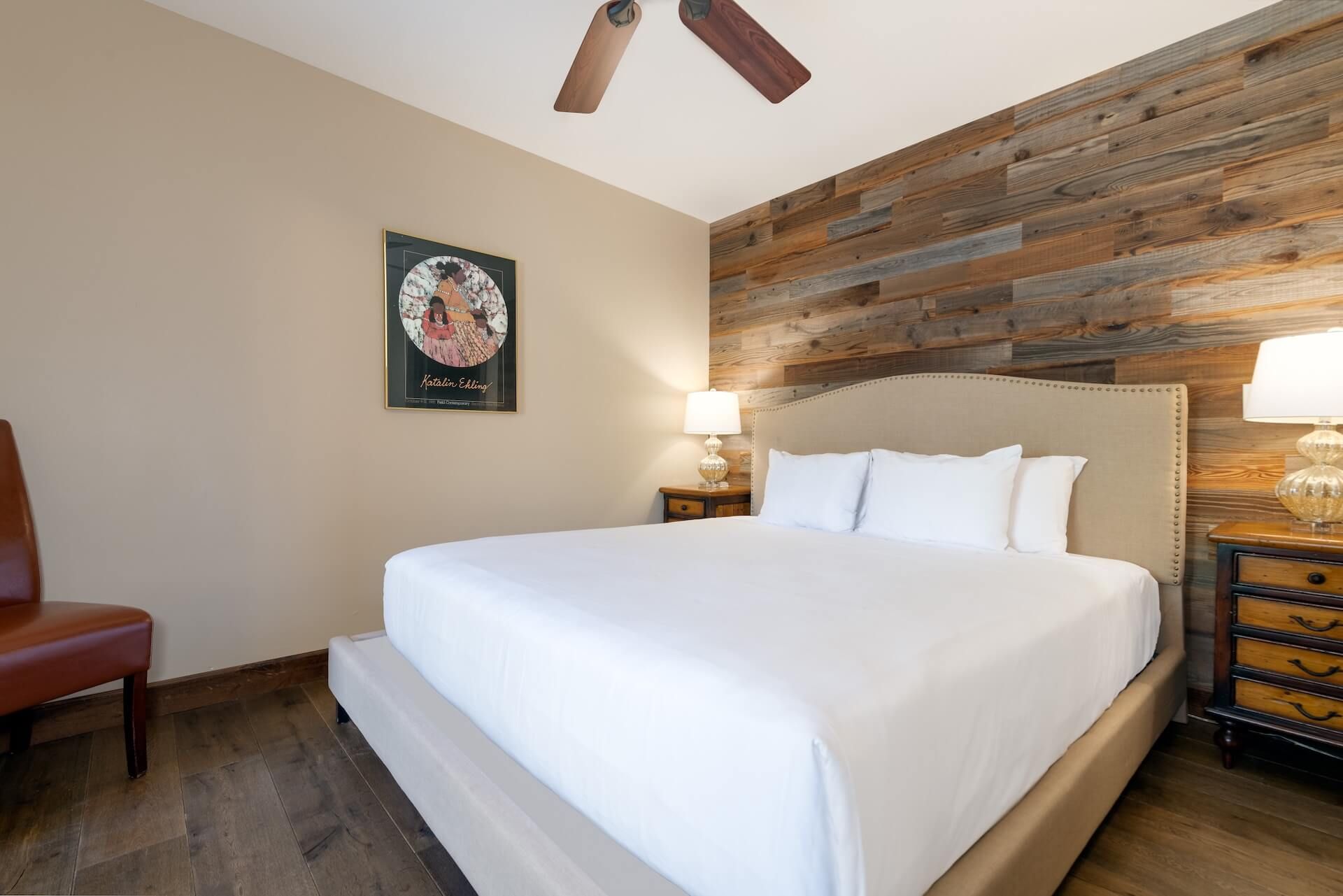 A hotel room with a large bed, a wooden accent wall, a framed picture on the wall, and a nightstand with a lamp.