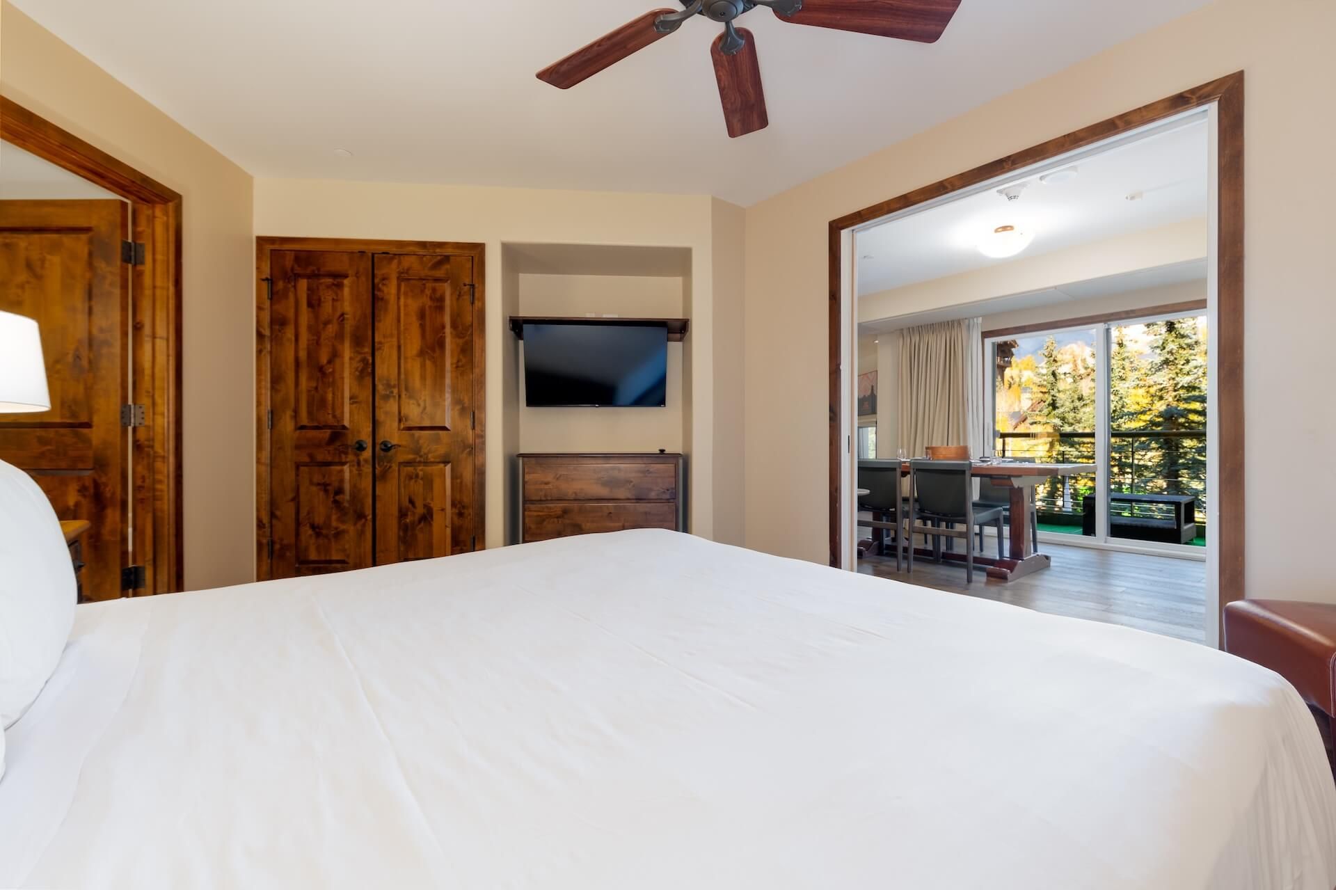 A bedroom with a large bed, wooden doors, a TV mounted on the wall, and a view of an outdoor patio area.