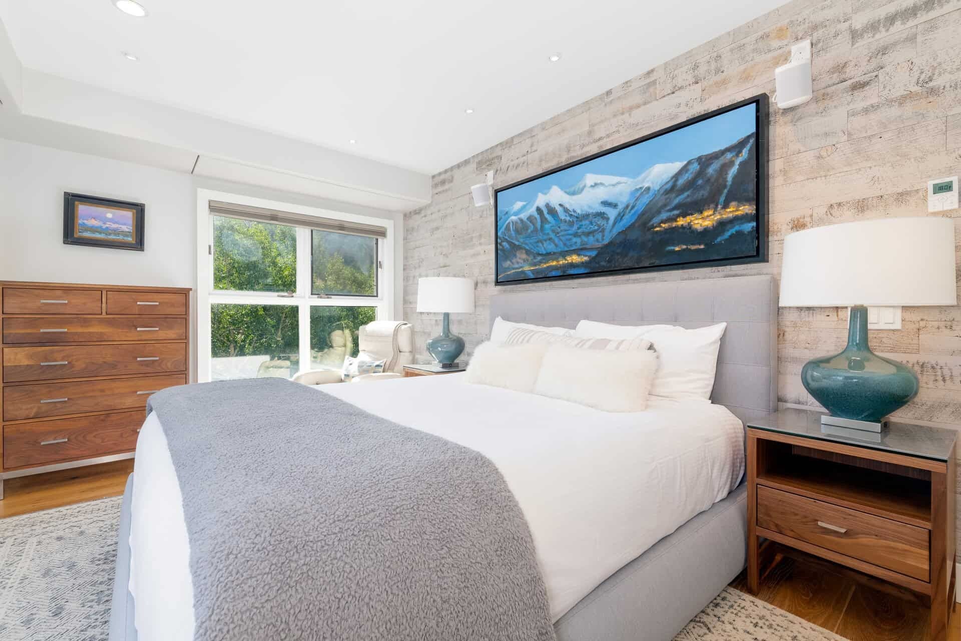 A bedroom with a large bed, a TV mounted on the wall, and a window with a view of greenery.