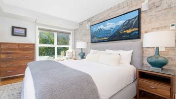 A bedroom with a large bed, a TV mounted on the wall, and a window with a view of greenery.