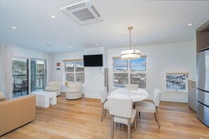 A modern living room with a round dining table, comfortable seating, a flat-screen TV mounted on the wall, and large windows offering a view of a snowy landscape. - Ski-In/Ski-Out 1BR Suite w/ Balcony Views + Pools (Mountain Village)