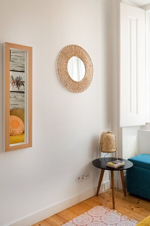 A room with a framed picture, a woven mirror, a wicker chair, a small table, and a blue sofa. - Graça Retreat | Spacious 2BR in Lisbon (Lisbon)
