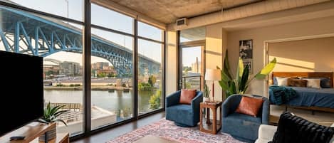 A modern living room with a large window overlooking a bridge and river, furnished with sofas, a coffee table, and a TV.