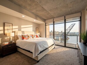 Ein Schlafzimmer mit einem großen Bett, einem Nachttisch, einer Kommode und Blick auf eine Brücke durch große Fenster.