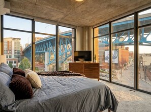 A bedroom with a large bed, a television on a wooden dresser, and a view of a blue bridge and cityscape through large windows. - Spacious 3-bedroom apartment with AC in soothing Cleveland (Cleveland)