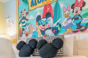 A room with a colorful wall mural featuring cartoon characters and a surfboard, a white couch, and three black Mickey Mouse ears pillows. - Luxury 7BR Disney Home • Pool Spa • Game Room -  Resort Windsor Island (Four Corners)