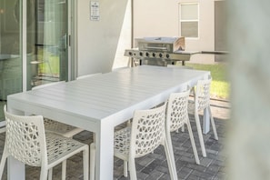 An outdoor patio with a white table and chairs, a grill, and a glass door. - Luxury 7BR Disney Home • Pool Spa • Game Room -  Resort Windsor Island (Four Corners)