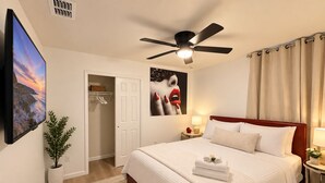 A hotel room with a bed, a ceiling fan, a nightstand, a TV, a door, a plant, and a painting. - New 5BR Home . Walk to Dining . Sleeps 10 (Jacksonville)