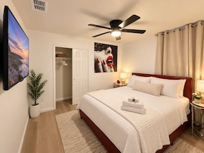 Habitación de hotel con cama, ventilador de techo, mesita de noche, televisor, puerta, planta y un cuadro.