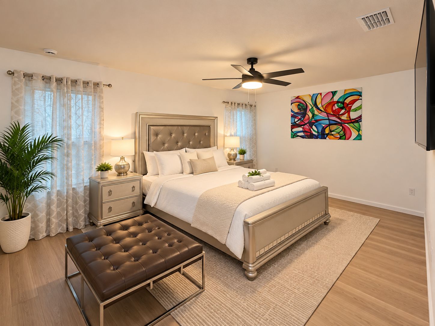 Une chambre à coucher comprenant un lit, des tables de chevet, un banc, un ventilateur de plafond et un tableau abstrait aux couleurs vives.