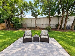 An outdoor patio area with two wicker chairs and a small table, surrounded by a brick floor and lush green grass. - New 5BR Home . Walk to Dining . Sleeps 10 (Jacksonville)