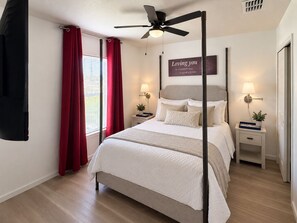 Un dormitorio con cama, ventilador de techo, mesita de noche con lámpara y ventana con cortinas.