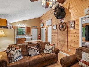 Een chaletinterieur met een houten wand, een bank met gestreepte kussens, een opgehangen dierenhoofd en een keukenhoek.