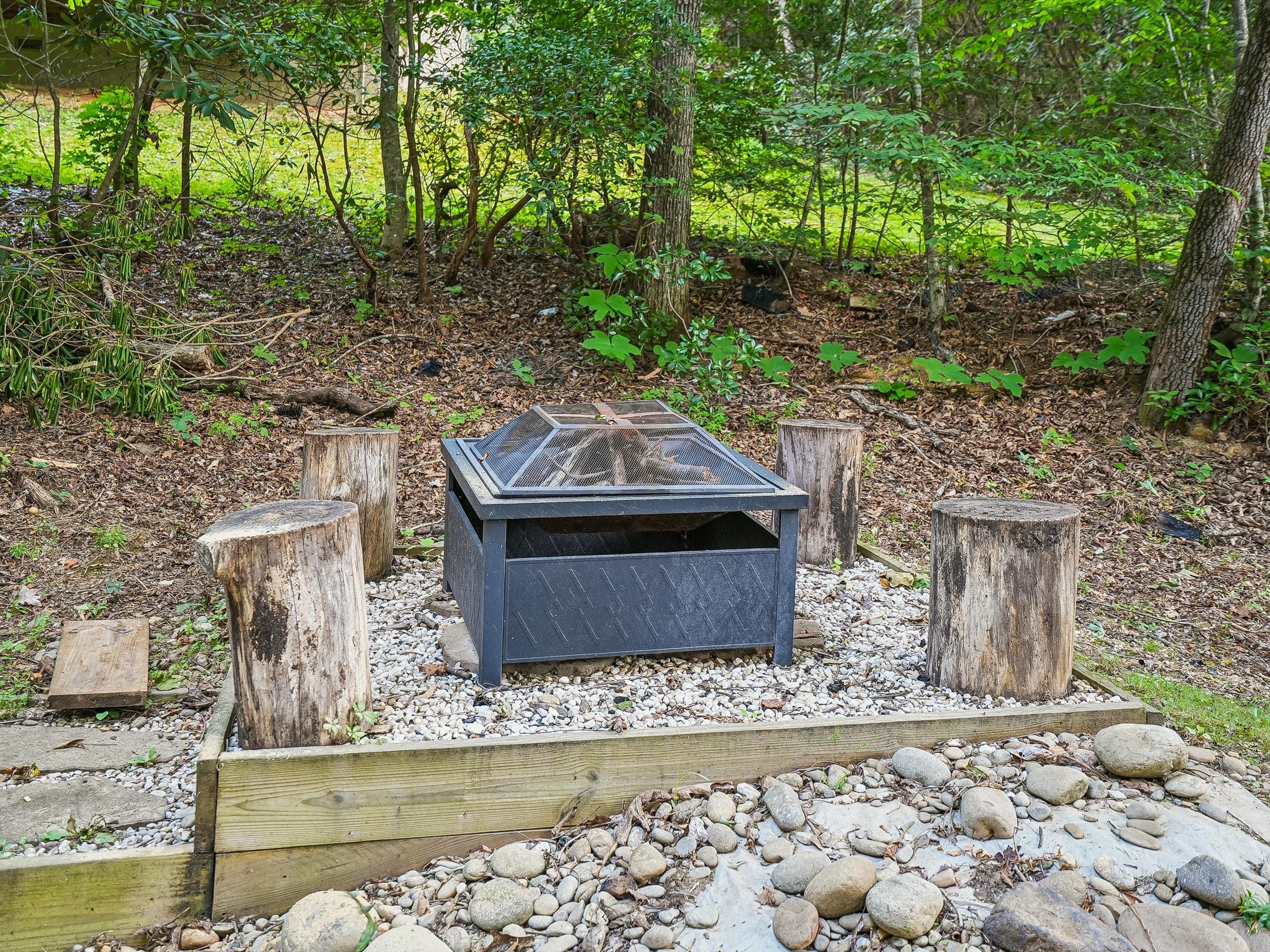 A fire pit with a metal grate in a forested area with wooden stumps and a gravel border.
