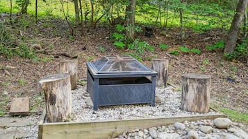 A fire pit with a metal grate in a forested area with wooden stumps and a gravel border.