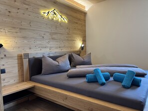 A modern bedroom with a wooden headboard, a bed with grey bedding, and blue towels.