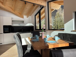 A modern kitchen with a wooden ceiling, a dining area with a wooden table and chairs, and large windows offering a view of trees and mountains.