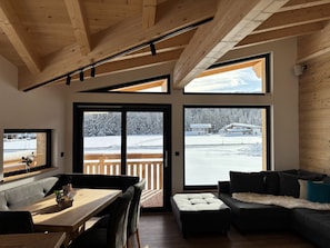 Un interior moderno con techo de madera, ventanas amplias, zona de comedor y área de estar con vistas a un paisaje nevado.