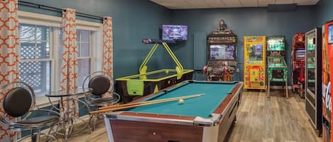 Uma mesa de bilhar com taco verde, duas cadeiras, uma televisão e jogos de fliperama.