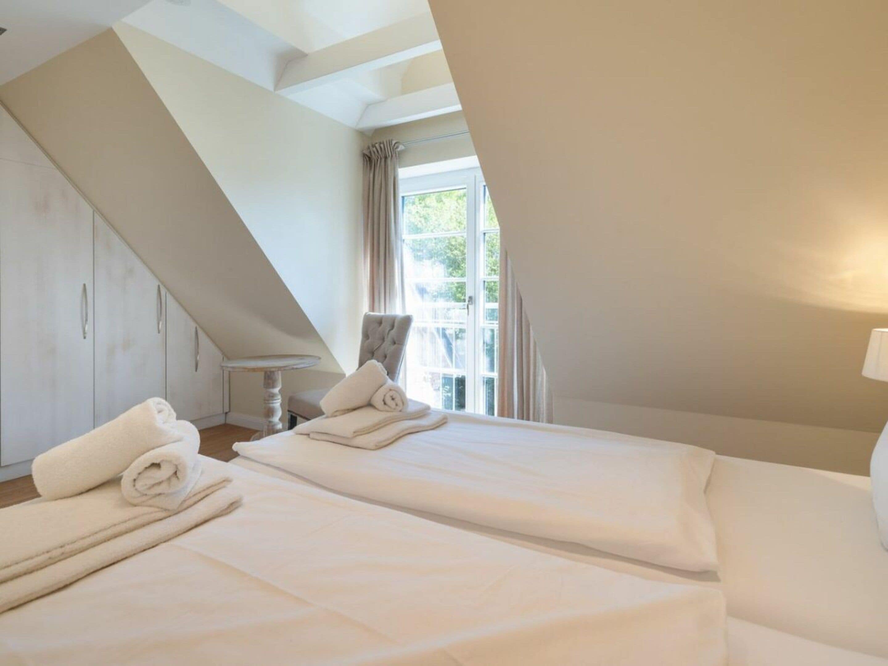 A bedroom with two beds, a chair, a table, and a window with curtains.