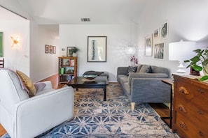 Una sala de estar con un sofá blanco, un sofá gris, una mesa de centro y un tapete estampado. Hay una estantería con libros y objetos decorativos, además de un cuadro en la pared.