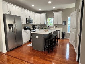 A modern kitchen with white cabinetry, stainless steel appliances, and a central island with stools. - The 414: Beach Retreat, Arcade, Fireplace, & Garage (Virginia Beach)
