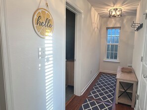 A hallway with a 'hello' sign, a patterned rug, a wooden floor, a window, and a small table with a basket. - The 414: Beach Retreat, Arcade, Fireplace, & Garage (Virginia Beach)