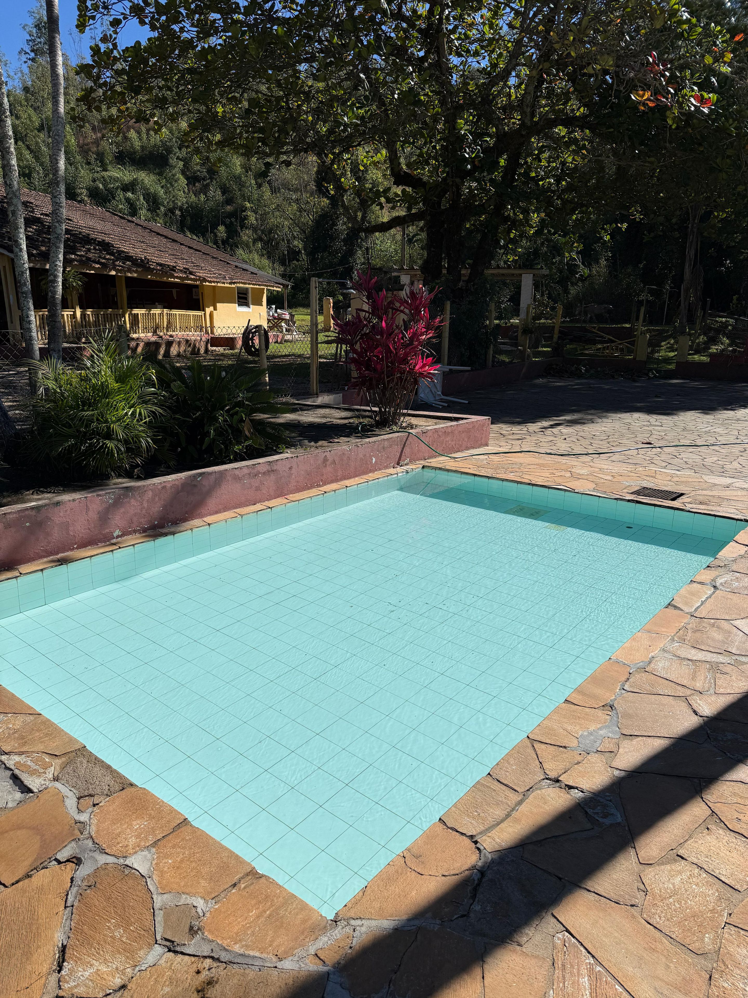 Una piscina con solárium de baldosas, bordeada de piedra, y un edificio amarillo al fondo.