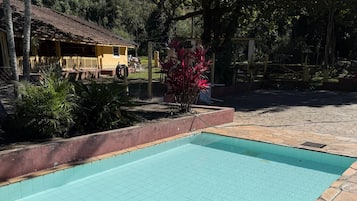 Una piscina con solárium de baldosas, bordeada de piedra, y un edificio amarillo al fondo.