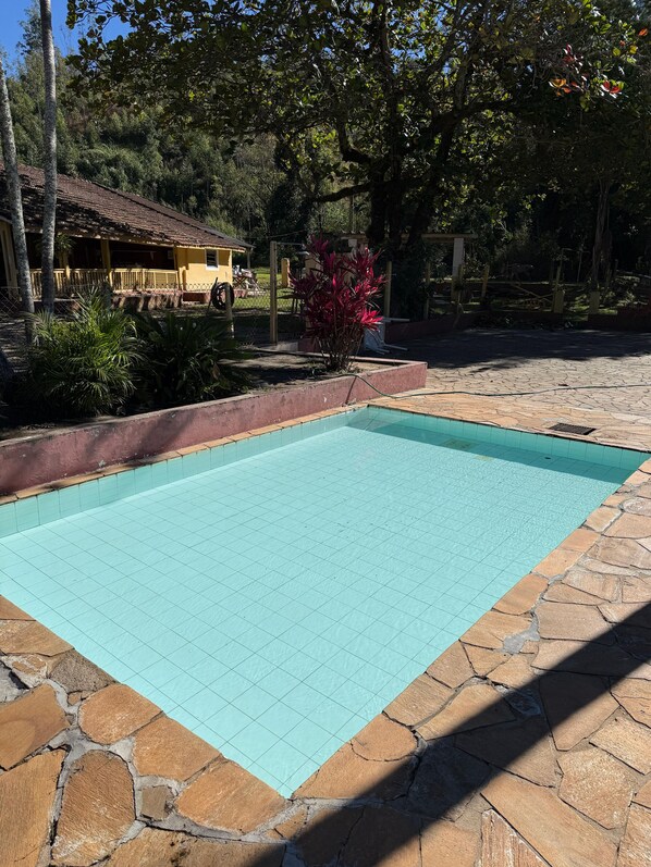Una piscina con solárium de baldosas, bordeada de piedra, y un edificio amarillo al fondo.