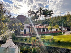 Un edificio residencial con techo de tejas rojas, rodeado de un césped cuidado y diversas especies de árboles, entre ellas palmeras.