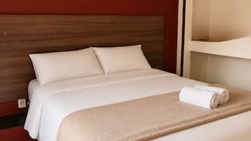 A hotel room with a bed, a headboard, two framed landscape paintings, a white towel on the bed, and a white shelf unit.