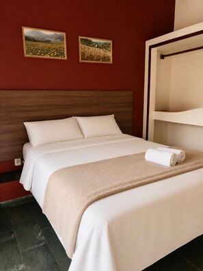 Habitación de hotel con una cama, cabecera, dos cuadros enmarcados de paisajes, una toalla blanca sobre la cama y un estante blanco.