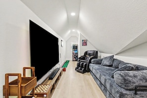 Un couloir étroit avec un téléviseur à écran plat, une table d’échecs en bois, un fauteuil massant et un canapé à motifs.