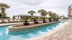 Piscina com espreguiçadeiras e plantas em vasos, cercada por palmeiras e um edifício ao fundo.