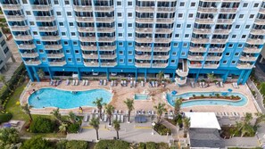 Um hotel com vários andares, com área de piscina, incluindo uma piscina grande central e uma piscina menor adjacente, rodeada por espreguiçadeiras e palmeiras.