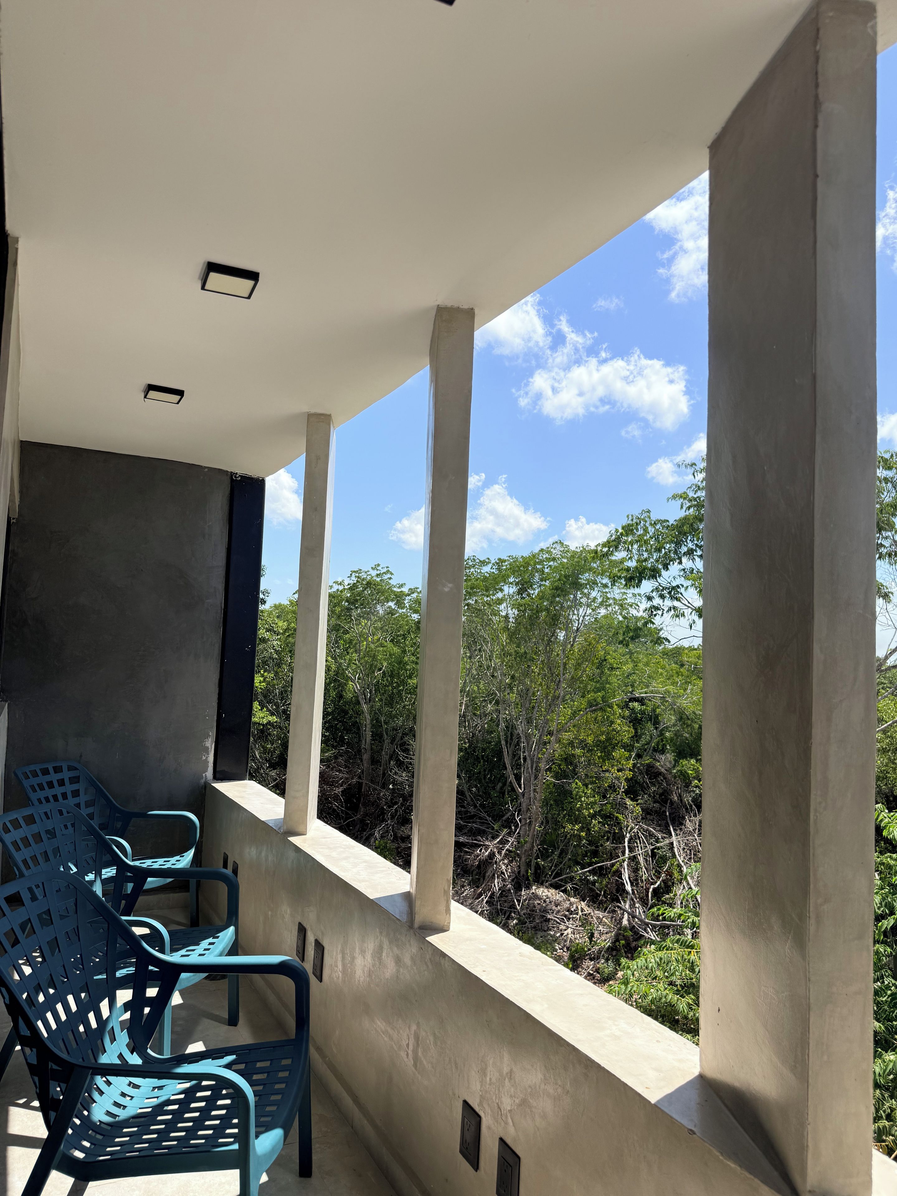Ein Balkon mit Blick auf Bäume und einen klaren Himmel. Blaue Stühle und eine weiße Decke sind vorhanden.
