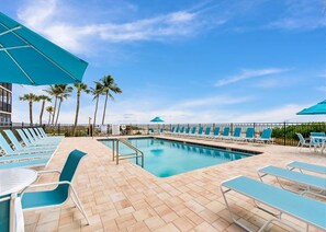 Un área junto a la piscina con sillones de descanso y sombrillas, palmeras y un cielo despejado.