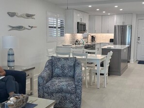 A modern kitchen with white cabinetry, a stainless steel refrigerator, and a dining area with white chairs and a patterned sofa. - Direct Gulf‑Front, 2/2 Corner Condo • Panoramic Beach Views & Steps to the Sand (Sanibel)