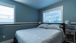 Un dormitorio con cama, dos ventanas con persianas, una mesita de noche con lámpara y un reloj de pared.