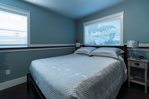 Quarto com cama, duas janelas com persianas, mesa de cabeceira com luminária e relógio na parede.