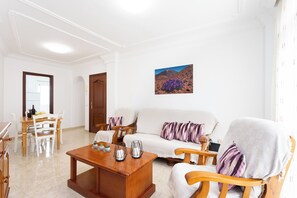 Ein Wohnzimmer mit Couch und Sesseln, einem Holztisch und einem Essbereich mit Tisch und Stühlen. Eine Tür führt in einen weiteren Raum.