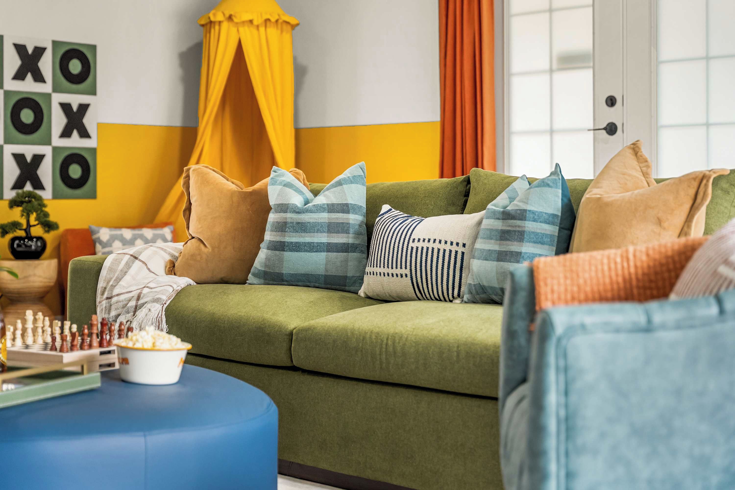 A green sofa with multiple cushions, a chessboard, and a popcorn bowl on a blue ottoman.