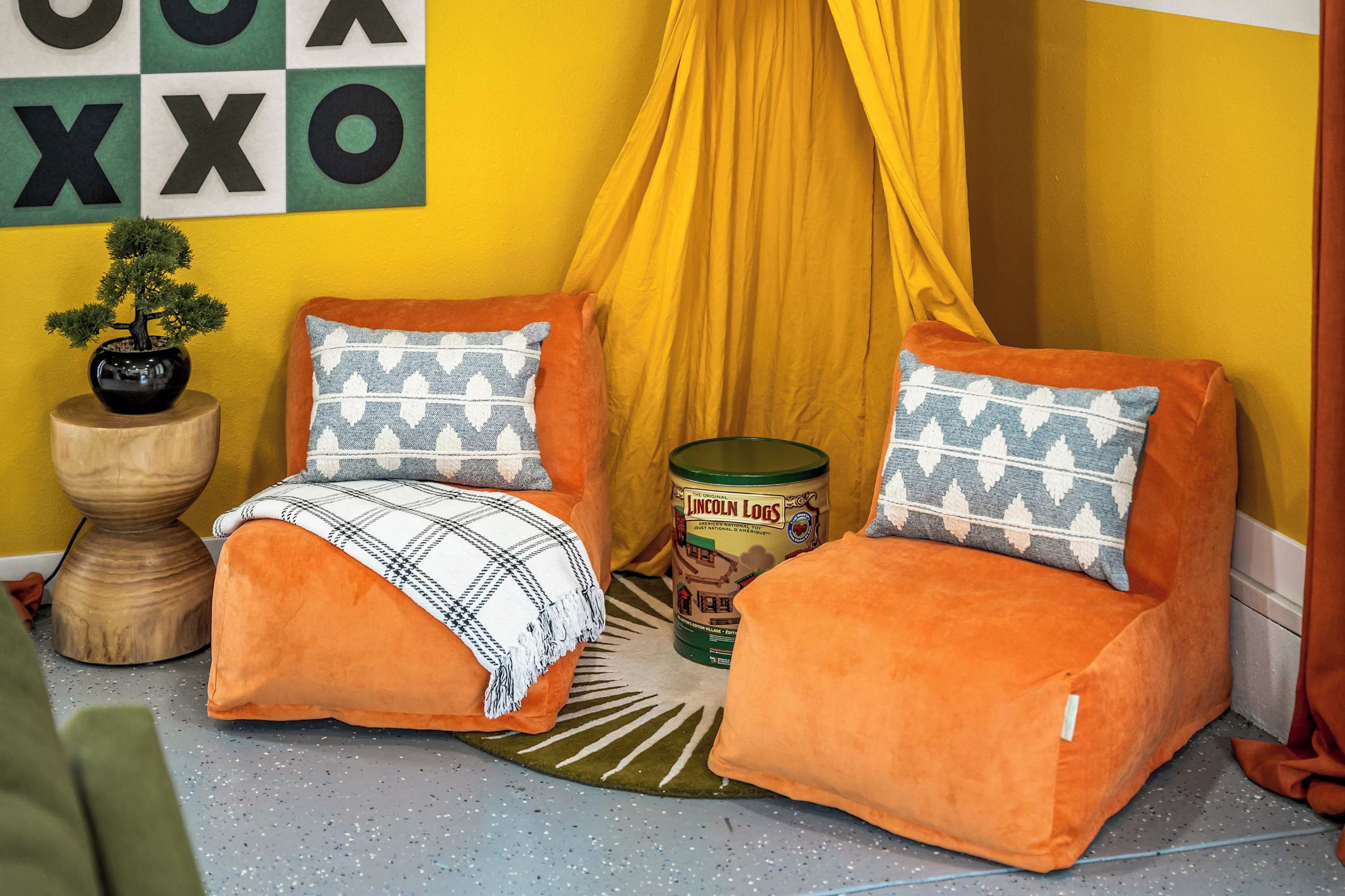 Un rincón acogedor con dos sillones naranja, una mesita de madera con un bonsái, una manta estampada y una lata de Lincoln Logs.