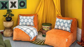 Un rincón acogedor con dos sillones naranja, una mesita de madera con un bonsái, una manta estampada y una lata de Lincoln Logs.