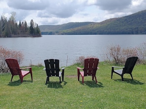 Three Adirondack chairs facing a lake with a forested hill in the background.