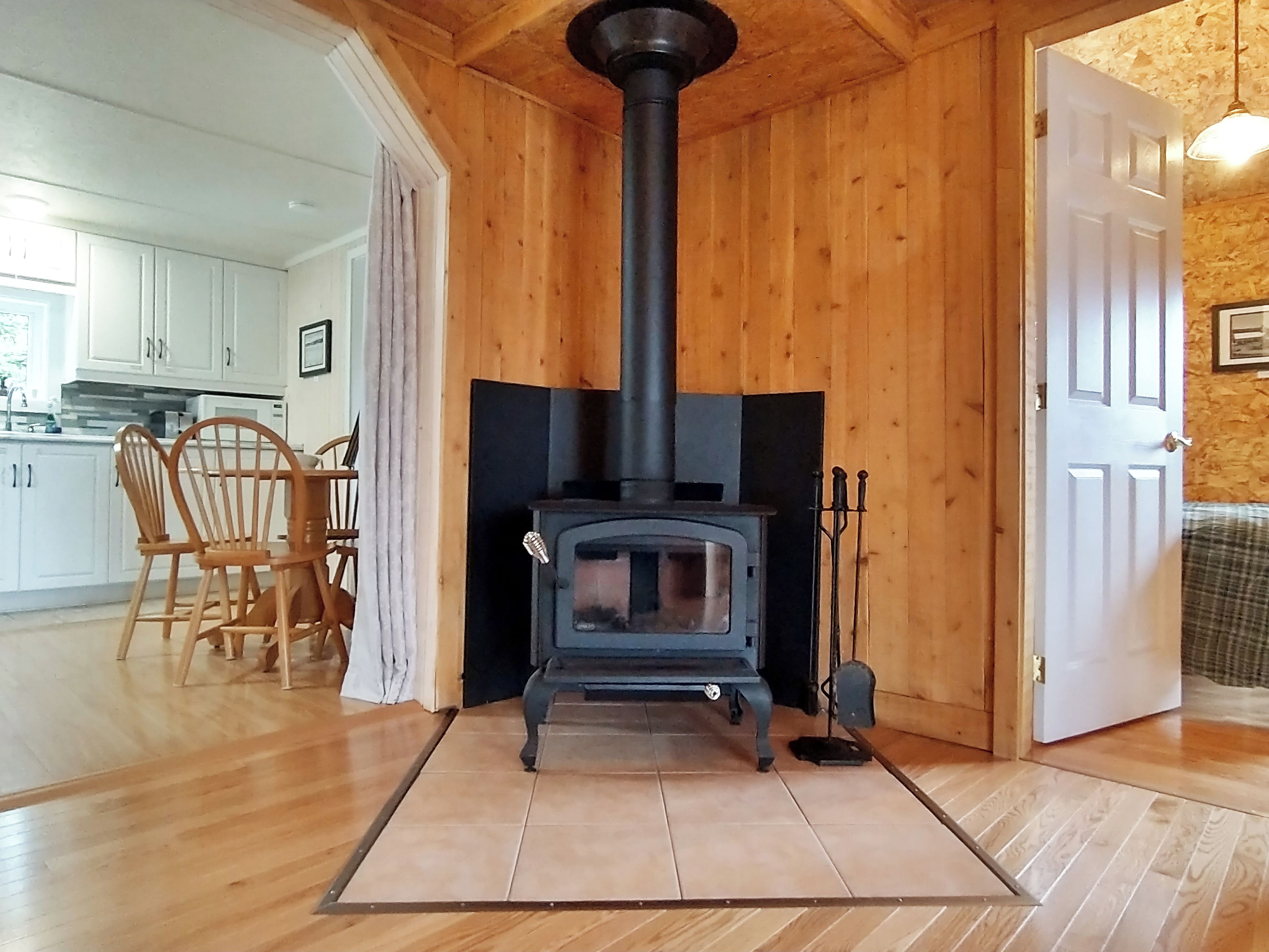 A room with a wood-burning stove, wooden flooring, a dining area with chairs, and a kitchen visible through an open door.