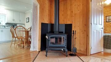A room with a wood-burning stove, wooden flooring, a dining area with chairs, and a kitchen visible through an open door.
