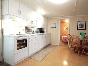 A compact kitchen with white cabinets, a built-in oven, and a dining area with a table and chairs. - Chalet Baptiste | Rustic cottage nestled in the mountains on scenic Lac Unique (Saint-François-de-Madawaska)