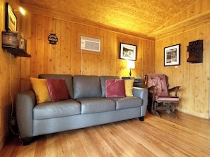 A wooden-paneled room with a grey sofa, a wooden chair, and a lamp. - Chalet Baptiste | Rustic cottage nestled in the mountains on scenic Lac Unique (Saint-François-de-Madawaska)